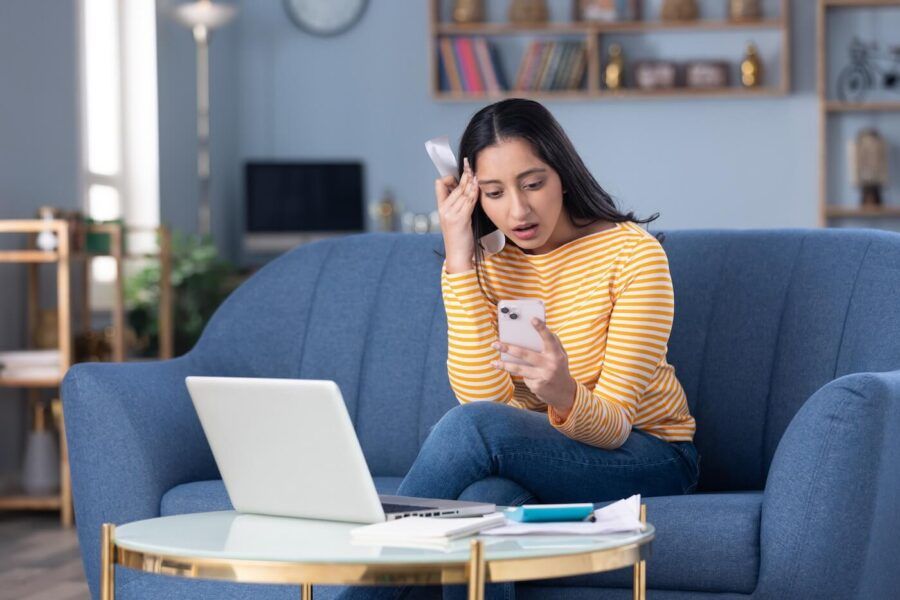 Concerned young woman seated on a blue sofa in a living room, looking at a smartphone while holding a receipt or bill. An open laptop, paperwork, and a notebook sit on a coffee table in front of the sofa, with bookshelves and home furnishings visible in the background.