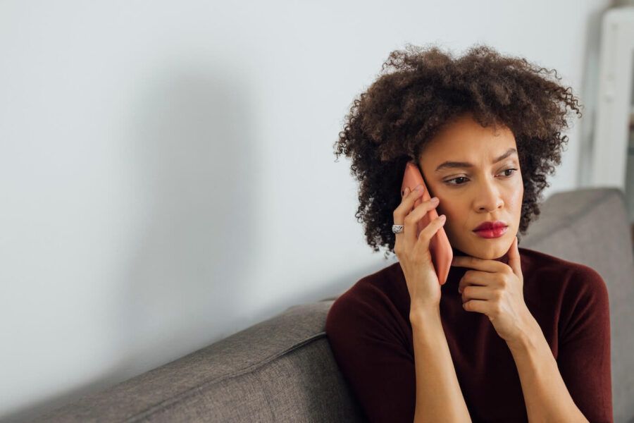 Concerned woman seated on a sofa in a home setting, holding a smartphone to one ear while looking off to the side. The image features a neutral-colored wall, soft natural lighting, and minimalist furnishings.