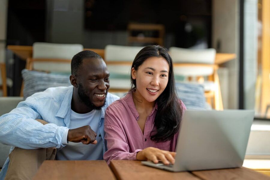 Happy couple sitting together in a living room, using a laptop on a coffee table, with a sofa, dining area, and large windows visible in the background of a modern home.