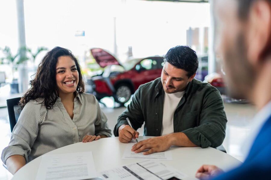 Happy couple seated at a desk in a car dealership, reviewing and signing paperwork with a sales representative. Documents are spread across the table, and vehicles are visible in the showroom background.