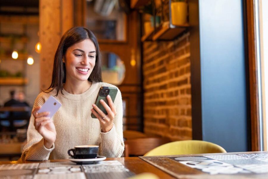 Smiling young woman sitting in a cozy cafe with a cup of coffee, checking her smartphone while holding a credit card