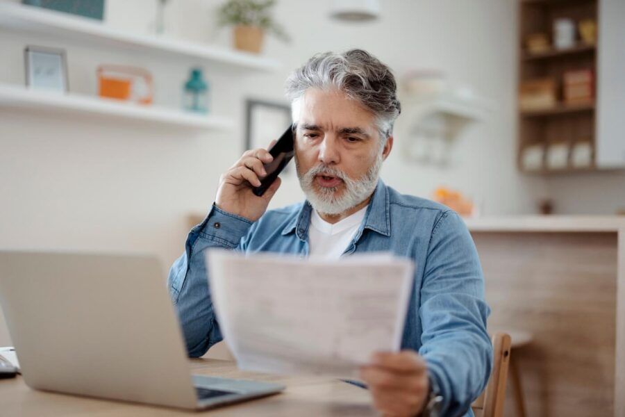 Focused senior man making a phone call while holding printed documents, with an open laptop on the desk