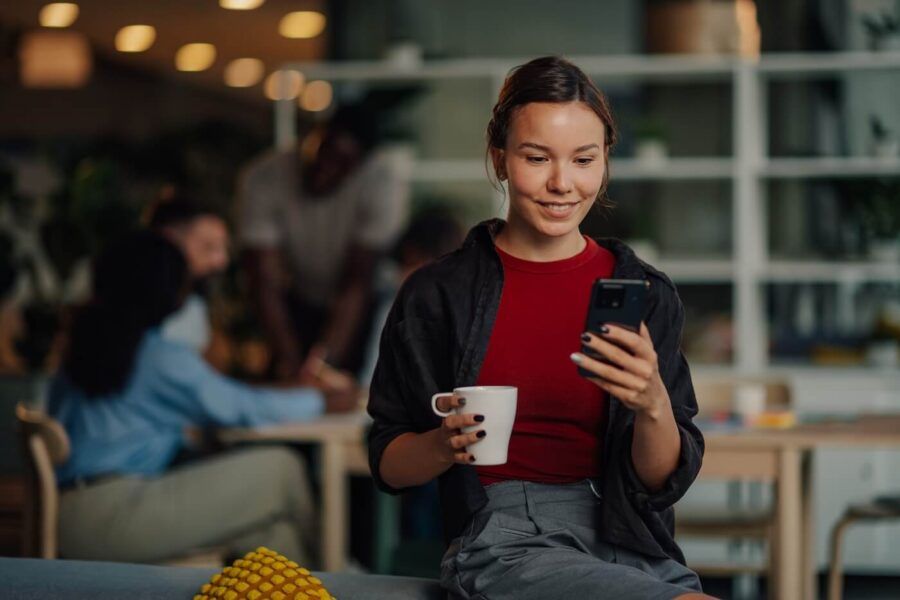 Smiling young woman on a sofa armrest using her phone and holding a mug in a busy café