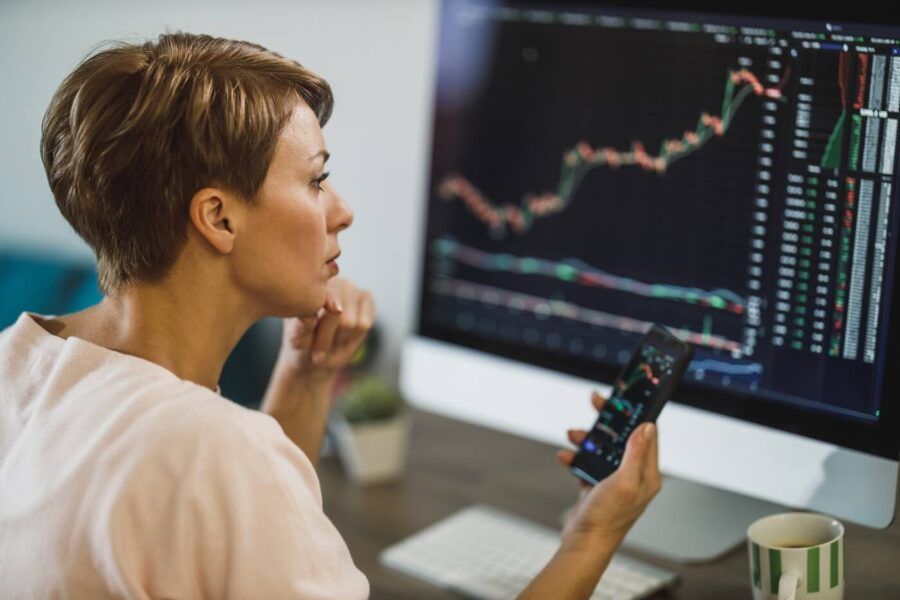 Focused woman sits at a desk holding a smartphone in one hand, with a desktop monitor showing line charts and market data, and a coffee mug nearby