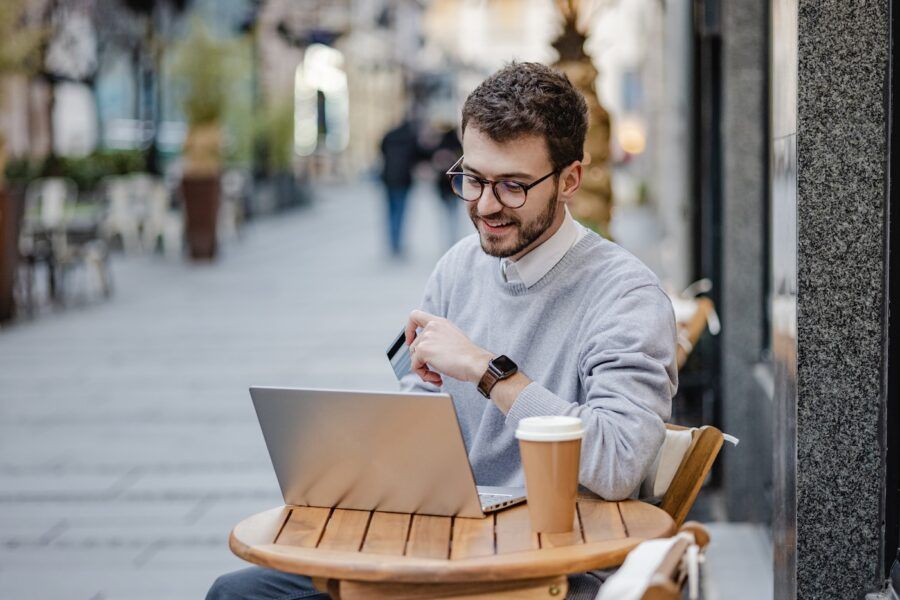 A young man using a laptop and holding a credit card while sitting at an outdoor cafe.