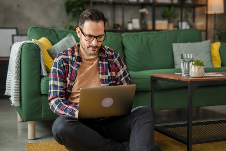Focused man using his laptop in the living room while sitting comfortably on the floor and leaning towards the green sofa