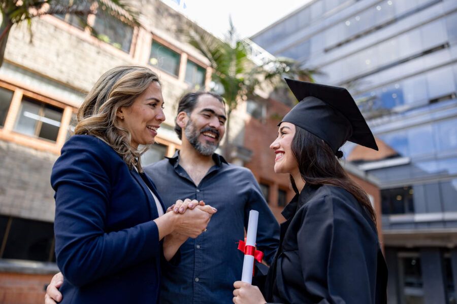 Smiling young woman wearing a black graduation cap and gown holds a rolled diploma with a red ribbon while shaking hands with her happy parents outdoors.