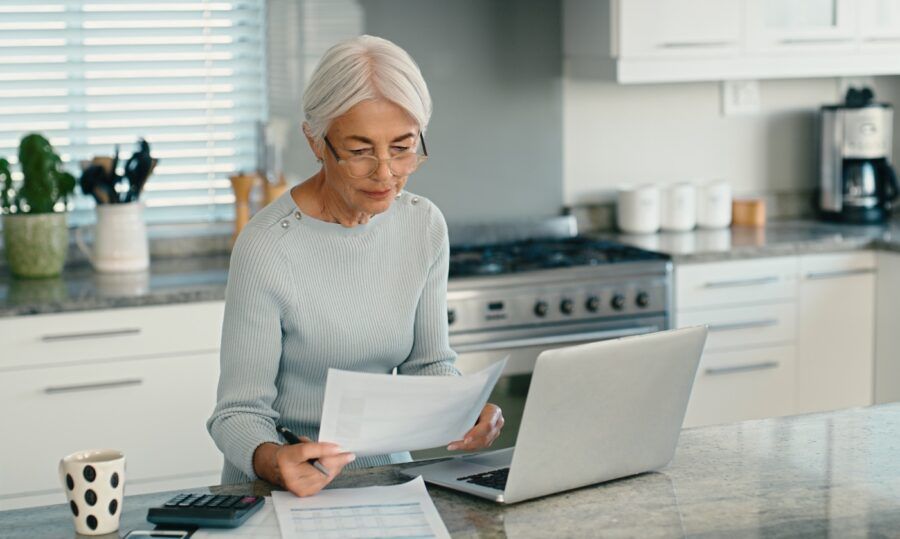 A senior woman looking at paper documents and working on her laptop in her kitchen.