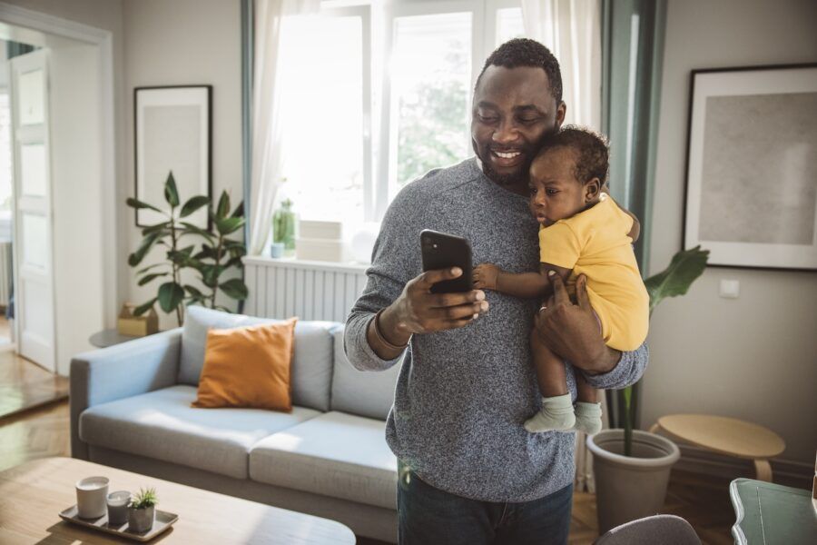 Young father at home holding his cute baby son in his arms, and using smart phone.