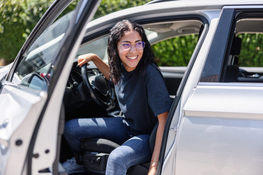 Happy young woman with glasses is stepping out of her parked car. The car door is open.
