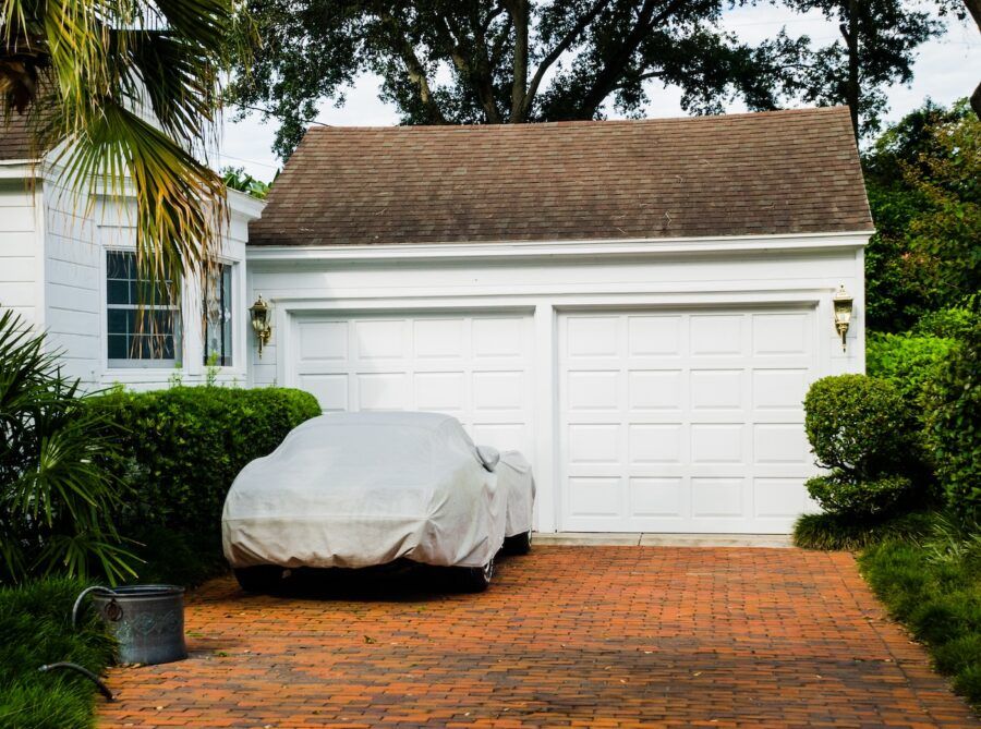 Covered car parked on brick driveway in front of white two car garage adjacent to house.
