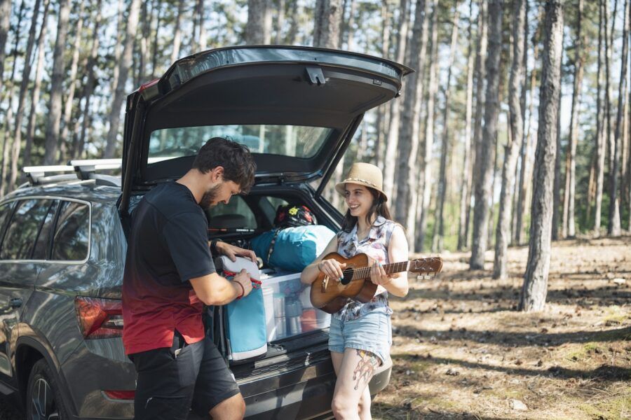 Young couple with their car packed for camping, woman is playing the guitar and man is listening.