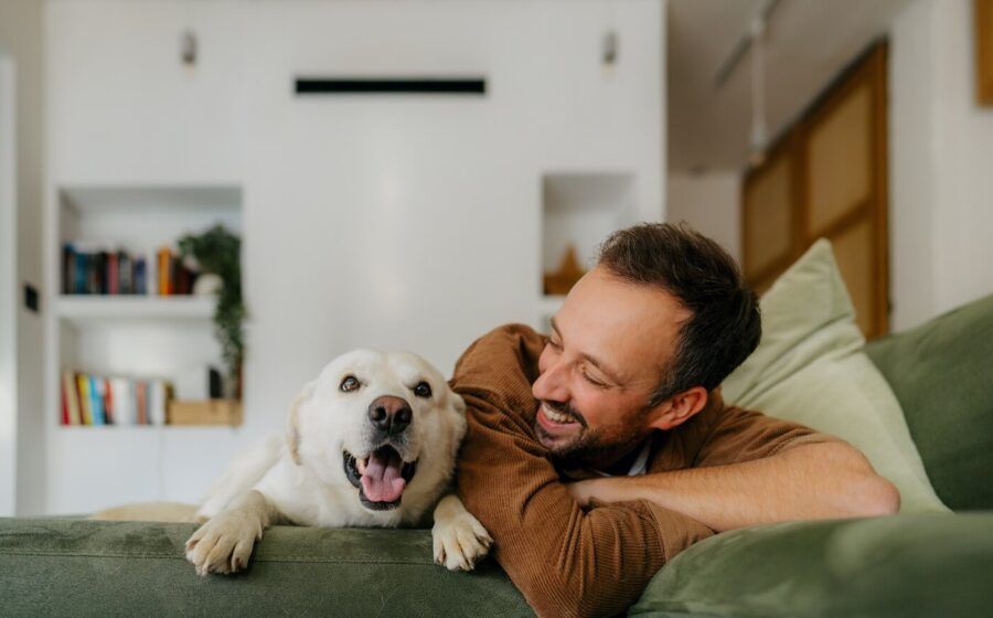 A smiling man relaxing on a couch with his Labrador lying beside him