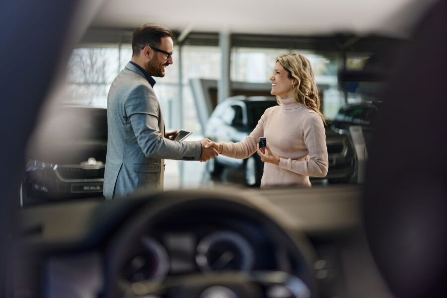 Happy salesman congratulating his female customer for leasing a new car in a showroom. The view is through a car windshield.
