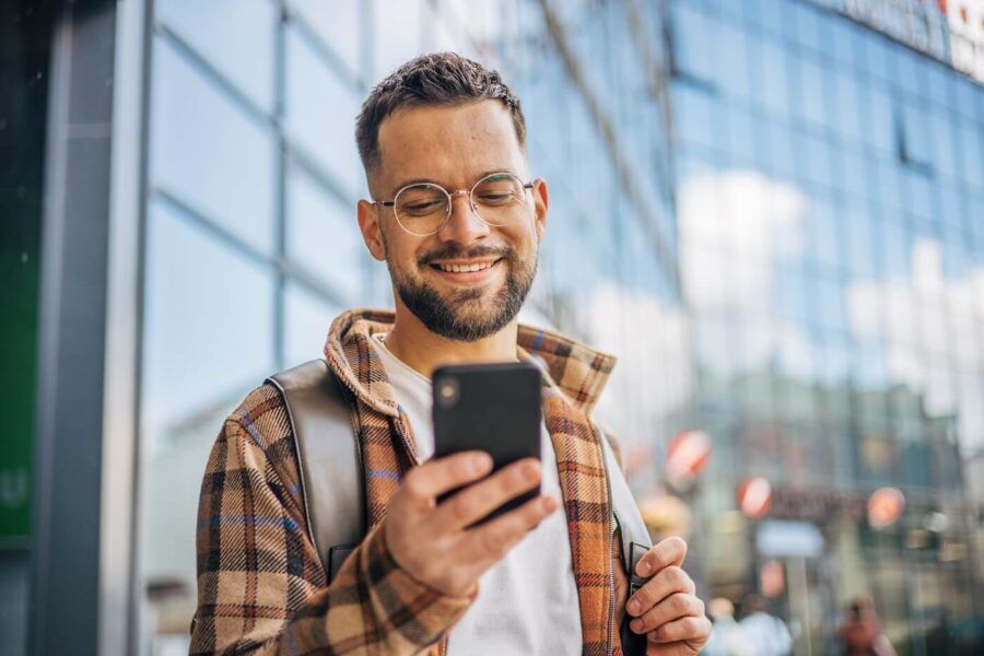 Smiling young man standing next to an office building and using his smartphone