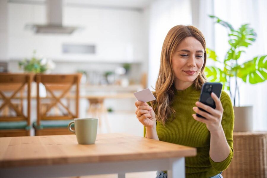 Smiling woman sitting at a kitchen table, holding a credit card and checking her smartphone, with a coffee mug beside her