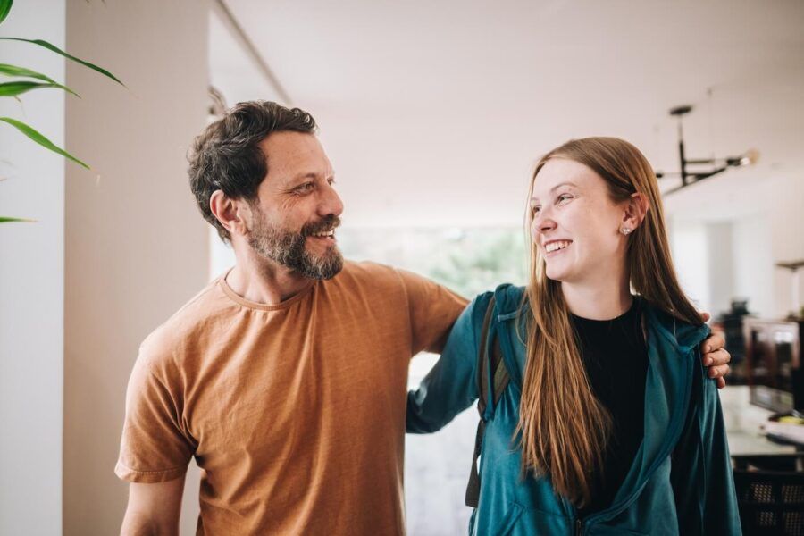 A father and a teenage daughter standing together in a bright home interior, with an arm around the teenager's shoulders and both looking toward each other near a large window.