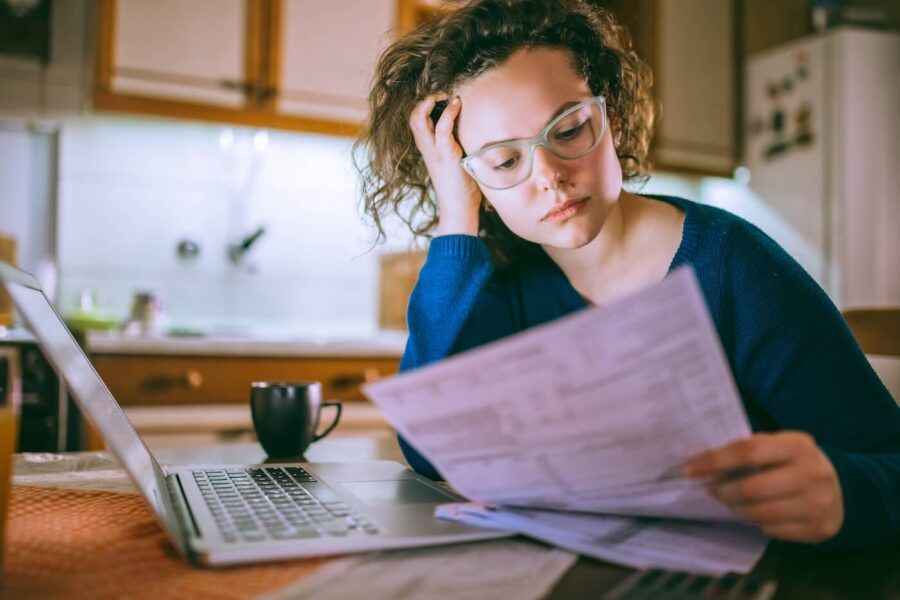 Concerned young woman reviewing her bills in the kitchen, a laptop and a mug standing on the table next to her.