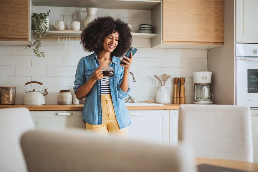 Smiling woman using her smartphone in the kitchen while holding a coffee mug
