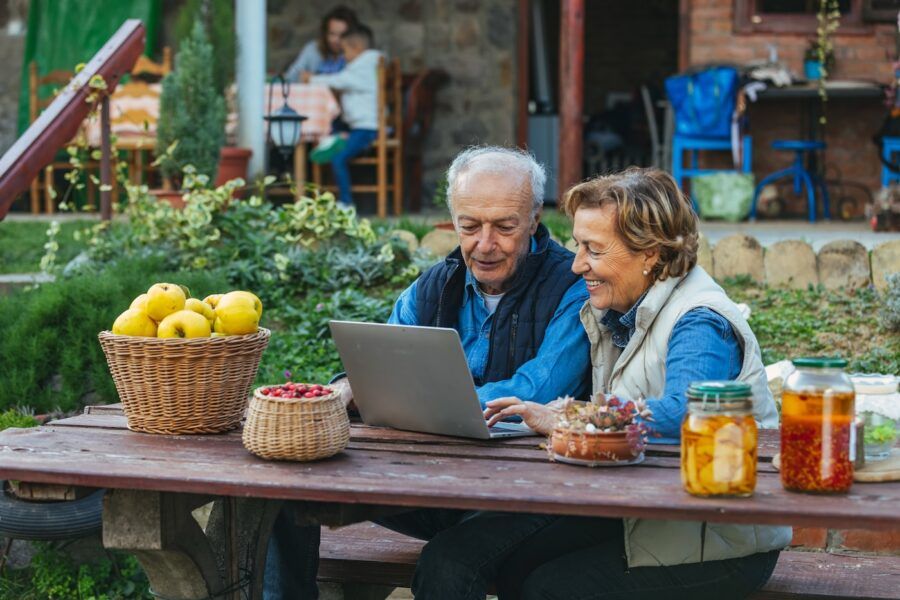 Senior couple using laptop computer to plan their finances while sitting at a picnic table in a backyard garden.