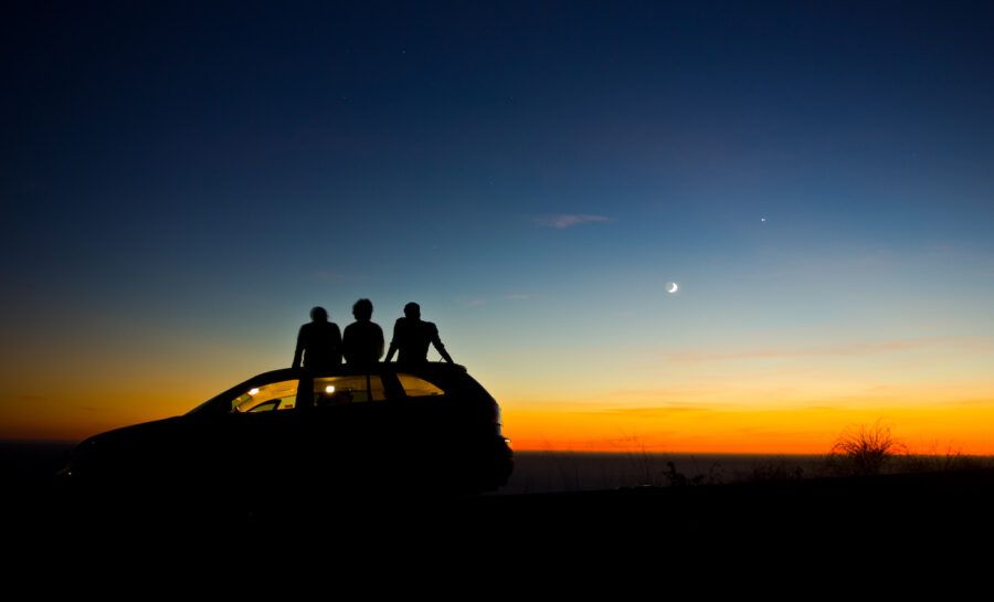 A silhouette from behind of three friends sitting on the roof of a car during sunset.