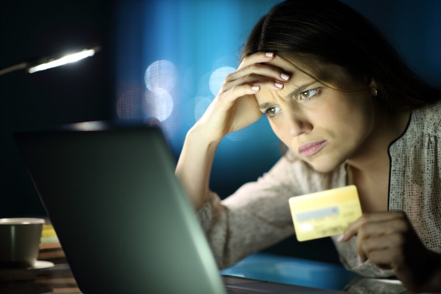 Frustrated woman holding her credit card stares at her laptop screen in the dark.