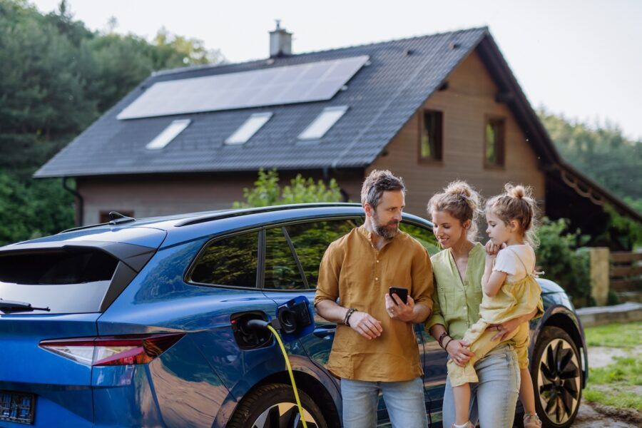 Two parents with their little daughter standing in front of their house with solar panels on the roof, comparing car insurance rates on a mobile phone while charging their electric car.