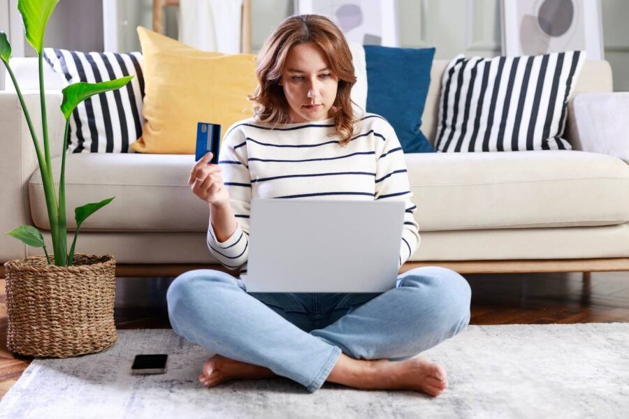 Woman sitting cross‑legged on the floor with a laptop on her lap, holding a credit card in her hand in a cozy living room