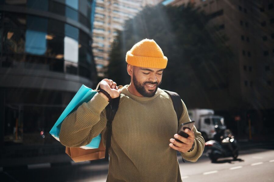 Smiling young man holding shopping bags and checking his phone outdoors on a sunny day