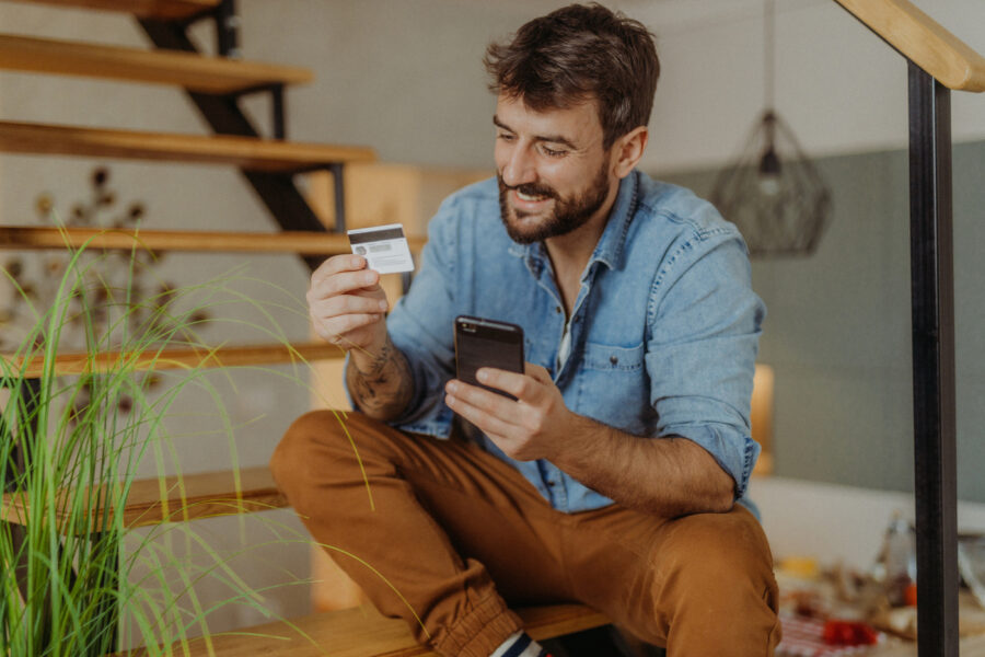 Young man sitting on the stairs using his credit card and checking his credit score on his mobile phone