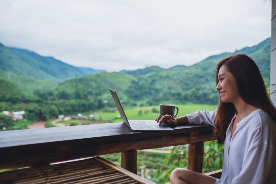 A woman working and typing on laptop computer while sitting on balcony with mountains and green nature background.