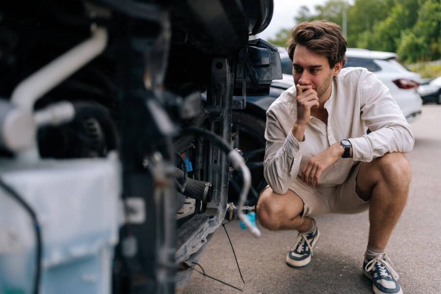 Stressed young driver man assesses damage to car following accident on street, concerned about expenses for repairs and insurance.