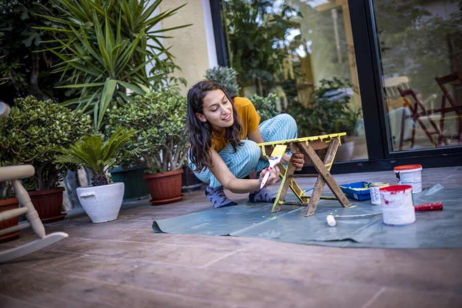 Young woman paints a small stool with yellow paint on her balcony.