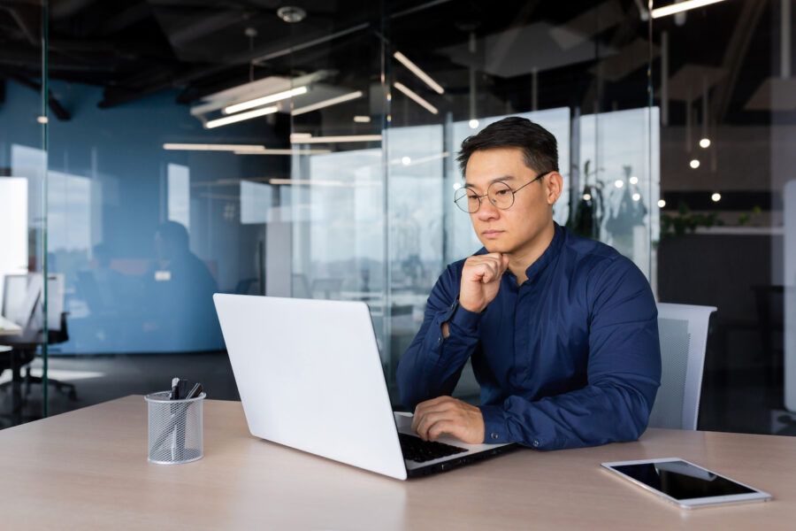 Serious thinking man working inside modern office, mature man in shirt and glasses using laptop at work, learning about how money market accounts are taxed.