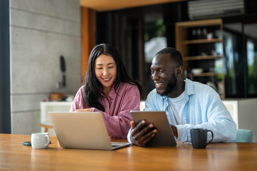 Smiling couple sitting at a table at home, using a laptop and tablet to plan their finances