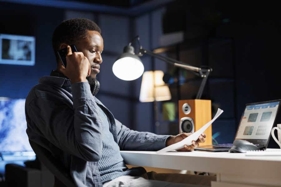 A man sitting at his desk at night, looking at a paper copy of his account balance and talking on the phone.
