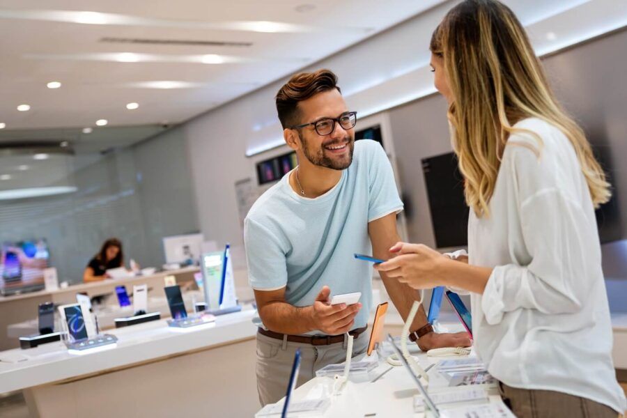 Happy couple stands at a display table in an electronics store, examining smartphones on stands while one holds a device and the other points with a pen, surrounded by other phones and retail displays.
