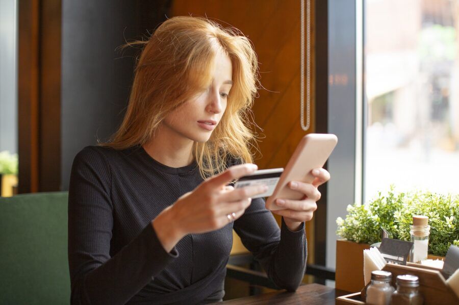 Red haired young woman making card payment through mobile phone to pay bills at a cafe.