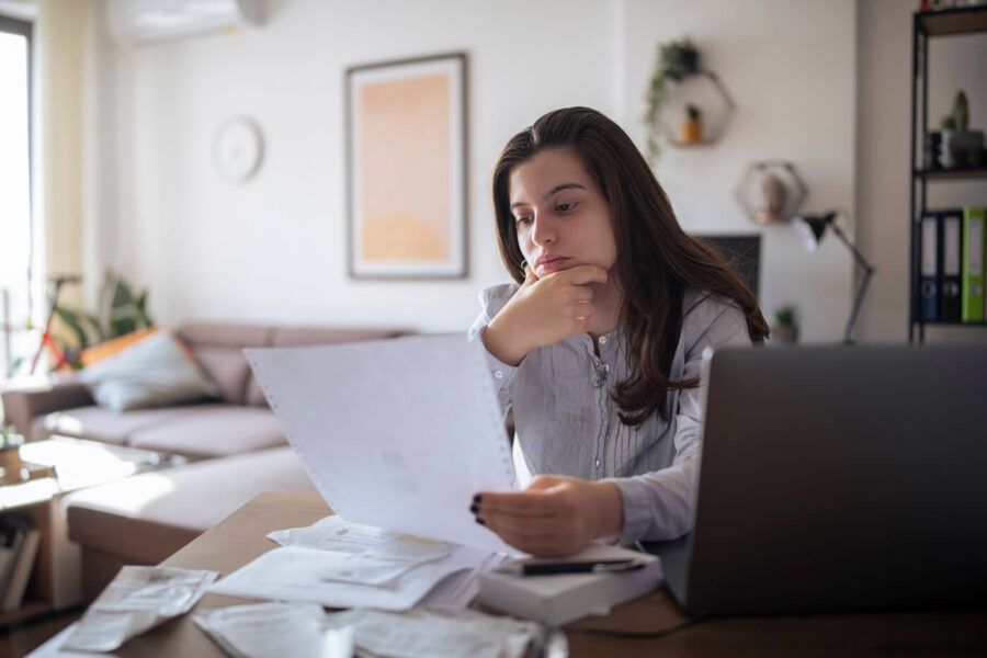 Pensive young woman reviewing a printed document while sitting at the desk with an open laptop and other printouts