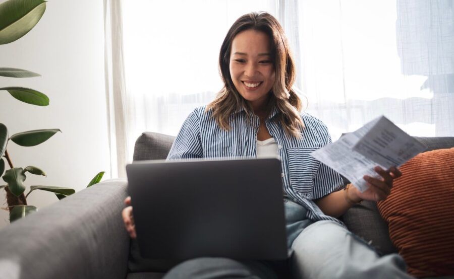 Smiling woman holding a letter and using her laptop while sitting on a sofa