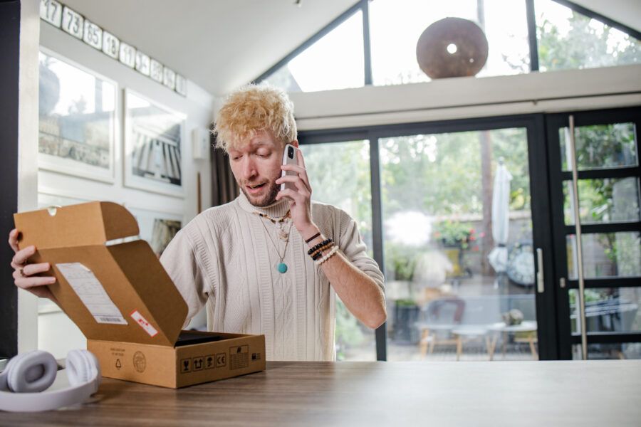 A young blond man talking on the phone while opening a package that has just arrived
