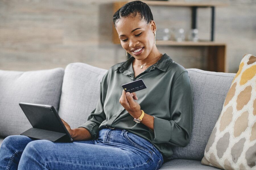 Woman holding debit card and shopping online with digital tablet on sofa at home, smiling and looking at card