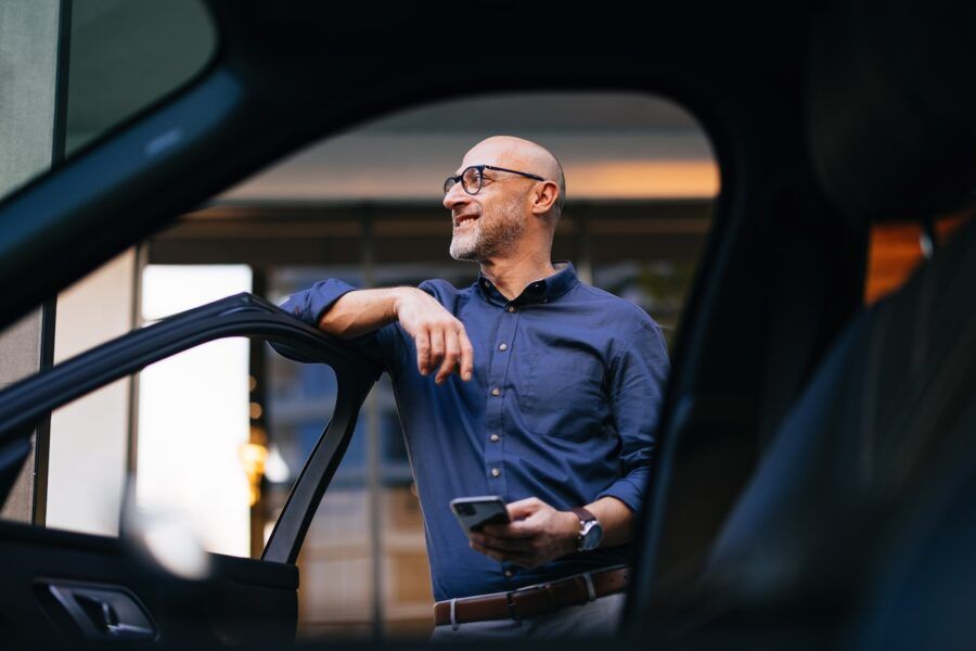 A mature businessman stands by his car with a smile, holding a smartphone in an urban setting.