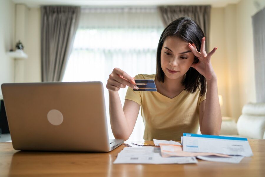 Stressed woman holding a credit card, sitting at her kitchen table in front of her laptop.