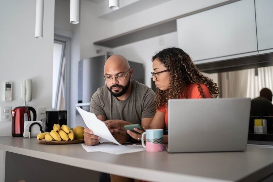 Focused couple reviewing their bills and using a laptop in the kitchen. There is a bunch of bananas, a kettle, and a mug on a countertop.