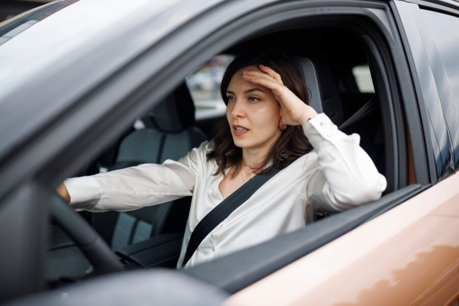 Tired woman sitting in the driver’s seat of a car with one hand resting on the steering wheel and the other near her head.