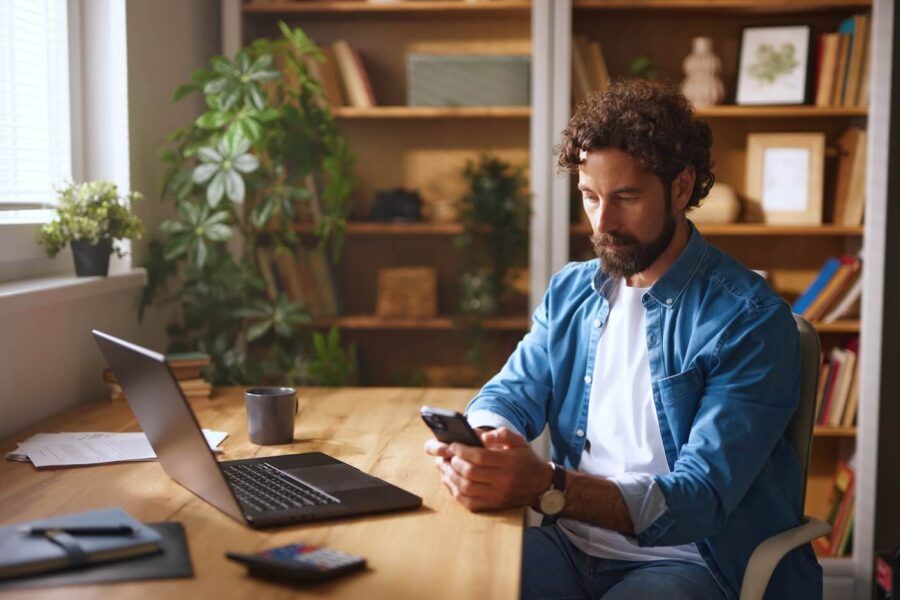 Focused man using his phone at the home office, with a laptop beside him
