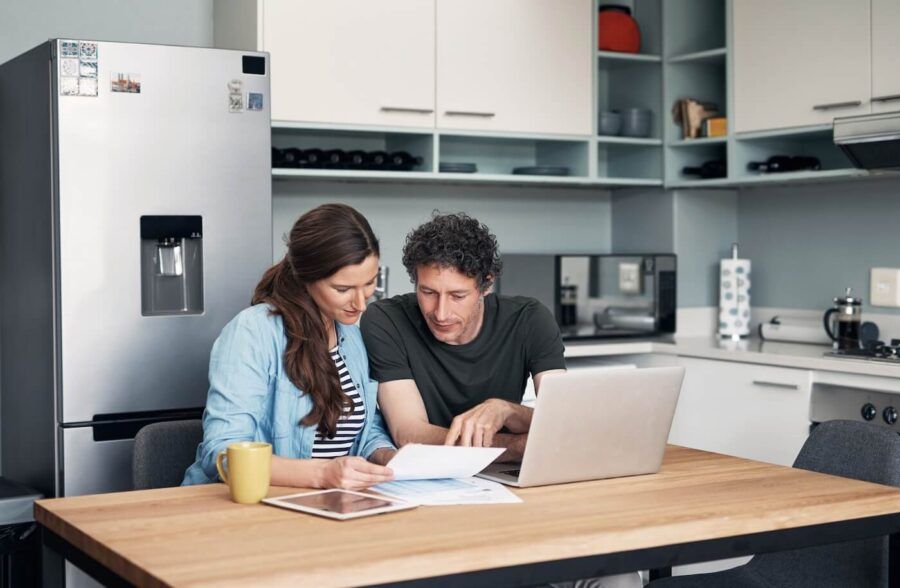 Mature couple reviewing printed documents in the kitchen, with a laptop and a mug beside them