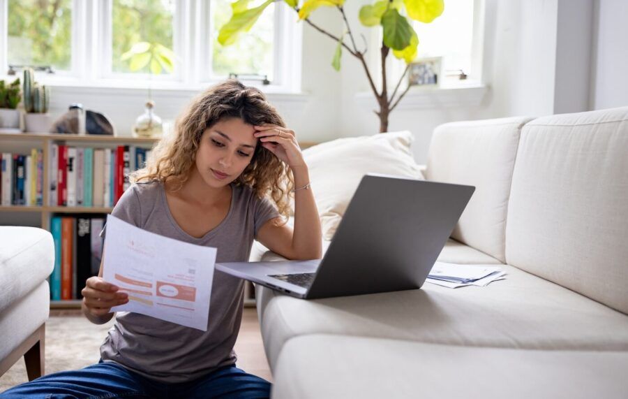 Woman seems worried about her home finances while reading her bank statement. She sits on the floor in her living room with a laptop on the sofa