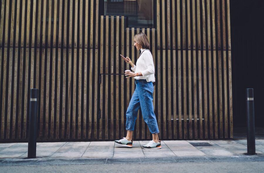 Side view of concentrated female pedestrian in stylish outfit checking credit report on mobile phone while walking along pavement near contemporary building